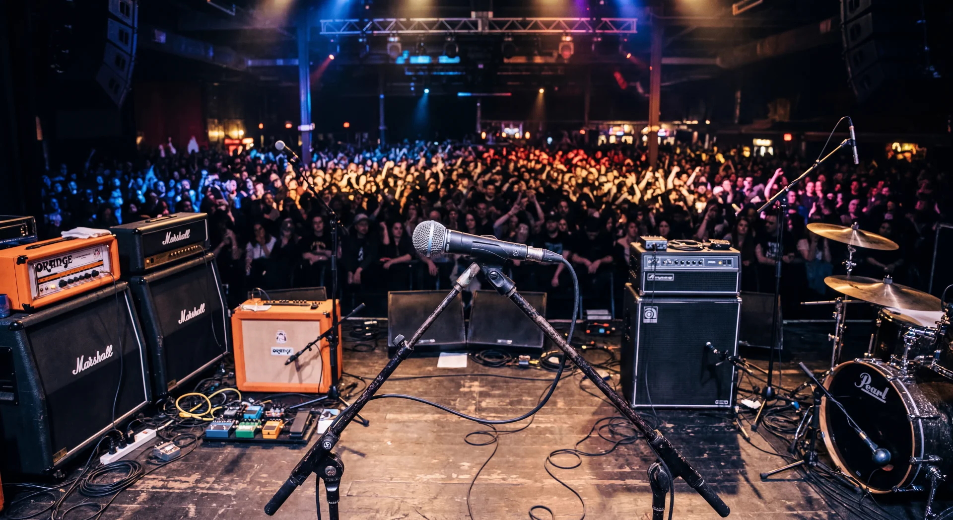 A packed crowd seen from the stage past a mic stand, amps and a drum kit